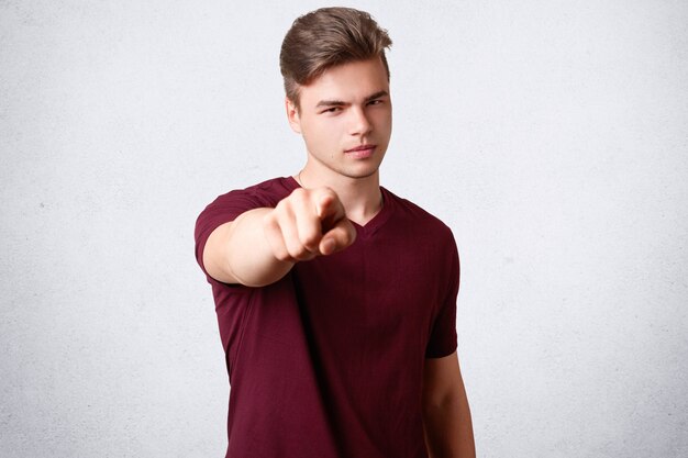 Serious male with trendy hairstyle, dressed in casual t shirt, points with fore finger, chooses you, poses on white concrete wall. I select you to help me with project work