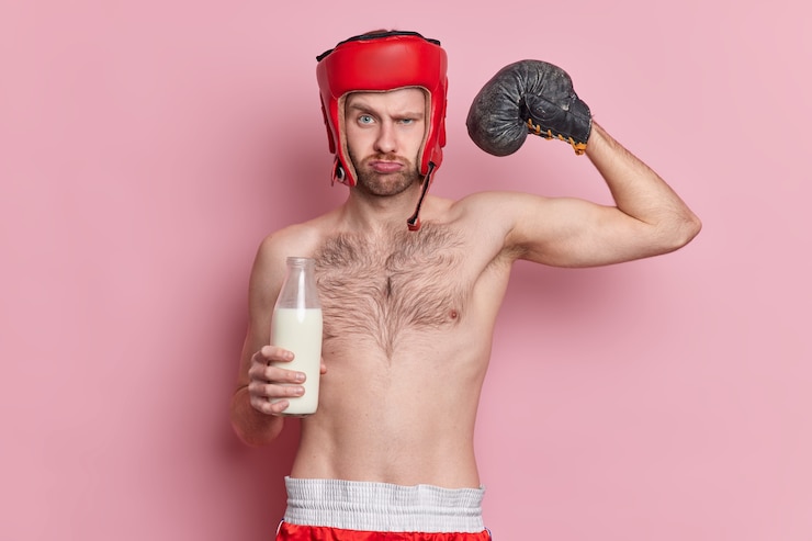 Free Photo serious man boxer with skinny body wears boxing gloves and hat raises arm shows muscles drinks milk for having strong biceps demonstrates his strength and power. sport and motivation concept