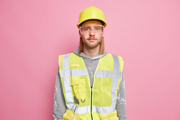 真面目な男性ビルダー エンジニアは、建設安全ヘルメットのユニフォーム グラスを着て、ピンクの壁越しに自信を持って仕事の準備ができているように見えます。自信を持っている職人または建設作業員