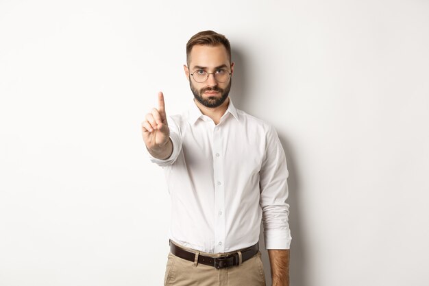 Serious man in glasses showing stop sign, shaking finger to prohibit and forbid, standing  white