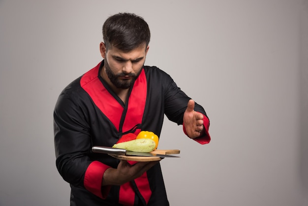 Серьезный человек, держащий деревянную доску с желтым перцем, цукини и ножом.