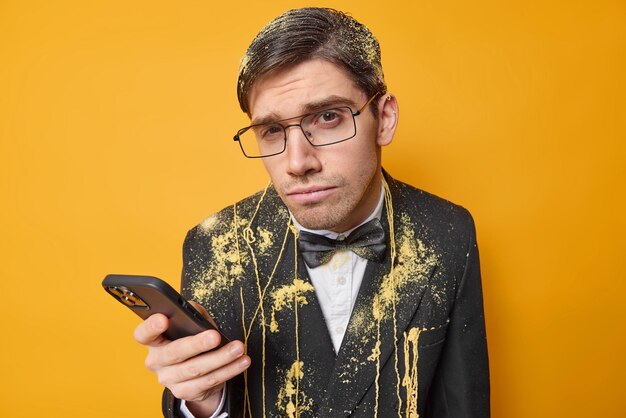 Серьезный мужчина смотрит с подозрением использует мобильный телефон для онлайн-чата, одетый в праздничную одежду, празднует особый случай, проводит время на вечеринке, изолированной на ярко-желтом фоне