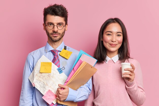 Serious man office worker holds folders wears formal shirt with attached stickers of reminding what to do. Pleased Asian woman drinks coffee helps groupmate with course work or startup project