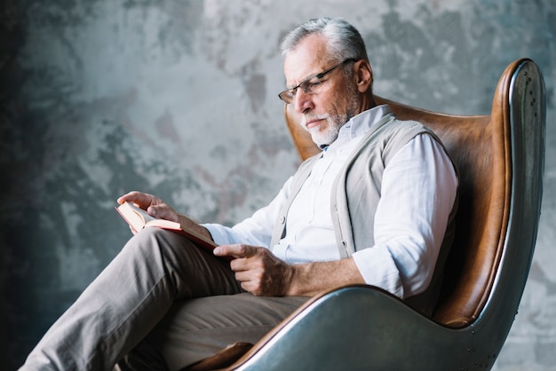 Serious man sitting on chair reading book against concrete wall