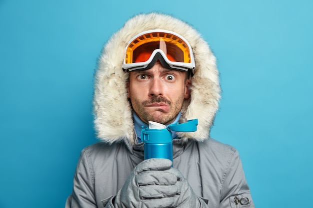 Серьезный мужчина дрожит от холода после катания на лыжах в морозный зимний день, держит фляжку с горячим напитком, носит лыжные очки и теплую куртку.