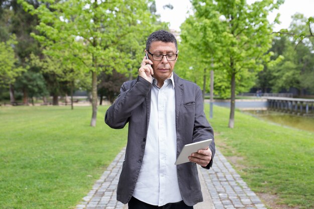 Серьезный человек с помощью планшета и разговаривает по телефону в парке