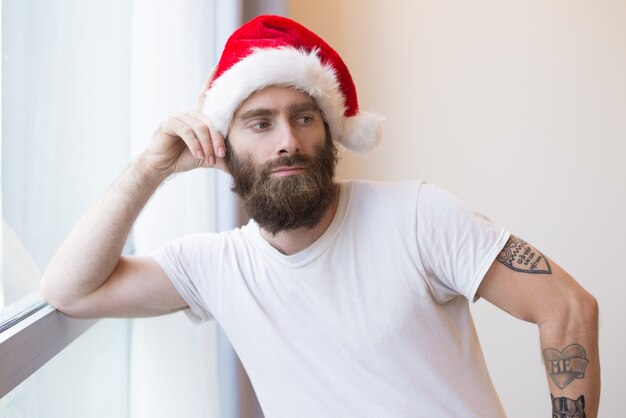 Serious man wearing Santa hat and leaning on window frame