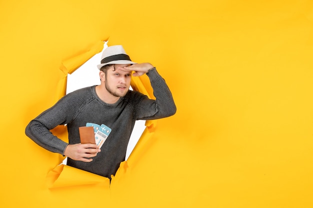 Бесплатное фото Серьезный мужчина в шляпе держит заграничный паспорт с билетом и сосредоточился на чем-то в разорванной на желтой стене