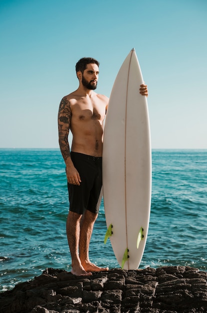 Serious man with surfboard standing on rock