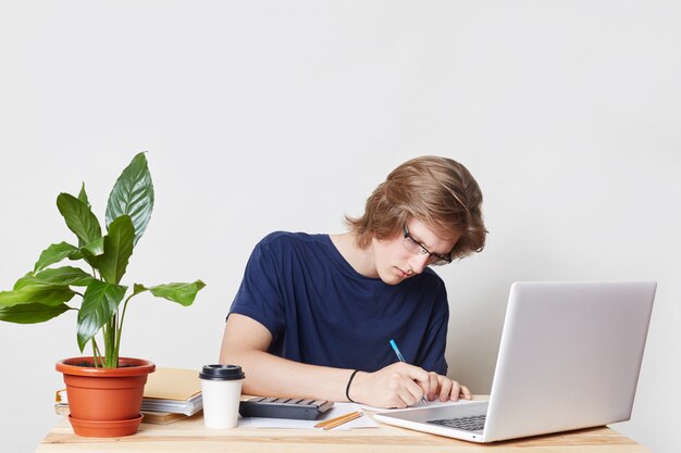 Serious man with trendy hairstyle, wears casual clothes, sits at work place, studies documents work on laptop computer