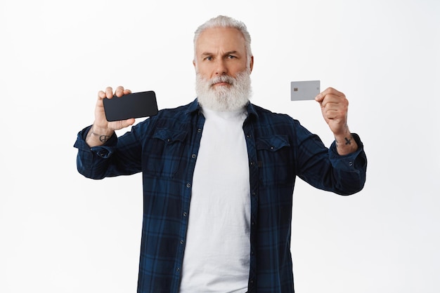 Uomo barbuto maturo serio che mostra lo schermo del telefono cellulare e la carta di credito che mostra qualcosa sul display dello smartphone in piedi su sfondo bianco spazio di copia
