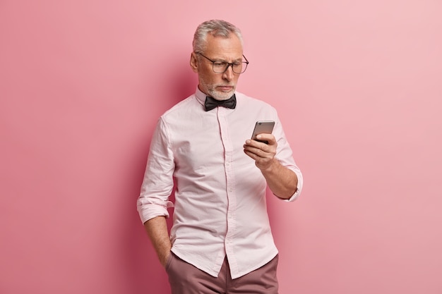 Serious mature man uses smartphone, reads news online, keeps hand in pocket, being always in touch, isolated over pink background.