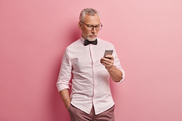 심각한 성숙한 남자는 스마트 폰을 사용하고, 온라인으로 뉴스를 읽고, 주머니에 손을 넣고, 항상 연락하고, 분홍색 배경 위에 절연되어 있습니다.