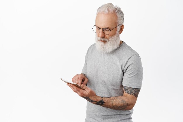 Серьезный взрослый мужчина с татуировками с помощью цифрового планшета в очках читает новости в социальных сетях, стоя на белом фоне