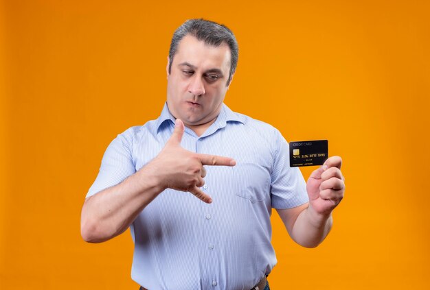 Serious middle age man in blue striped shirt looking at credit card on an orange background