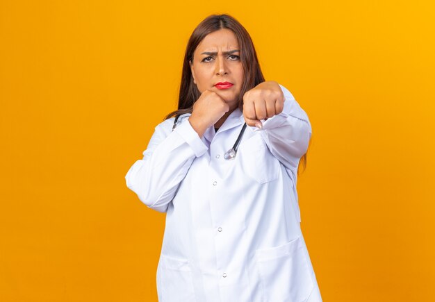 Серьезная женщина-врач среднего возраста в белом халате со стетоскопом позирует, как боксер со сжатым кулаком