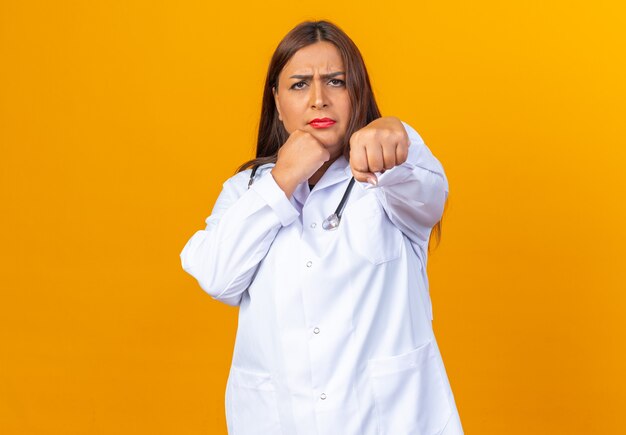 Serious middle age woman doctor in white coat with stethoscope posing like a boxer with clenched fist