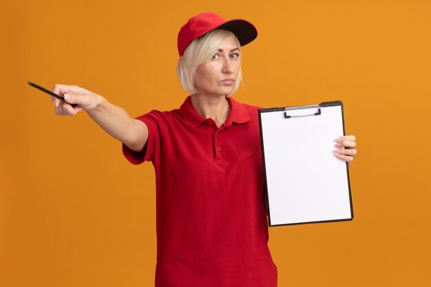 Free photo serious middle-aged blonde delivery woman in red uniform and cap holding clipboard looking at front pointing at side with pencil isolated on orange wall