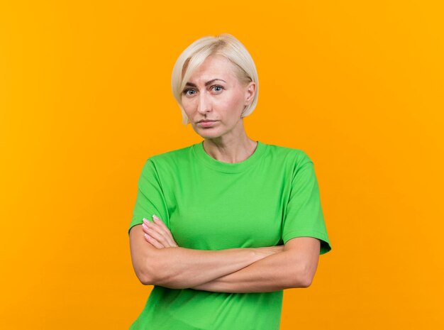 Serious middle-aged blonde slavic woman standing with closed posture looking at front isolated on yellow wall with copy space