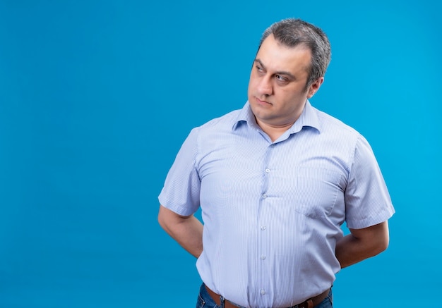 Serious middle-aged man in blue striped shirt thinking put hands on back on a blue background