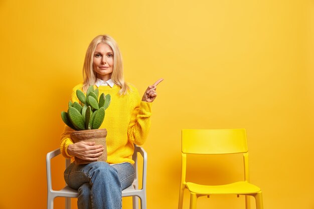 Serious middle aged woman poses with cactus at chair looks confidently and points away on copy space