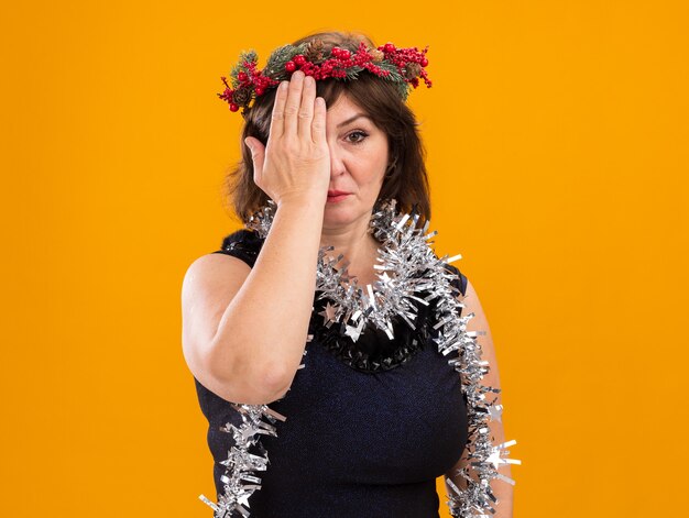 Serious middle-aged woman wearing christmas head wreath and tinsel garland around neck looking at camera covering half of face with hand isolated on orange background