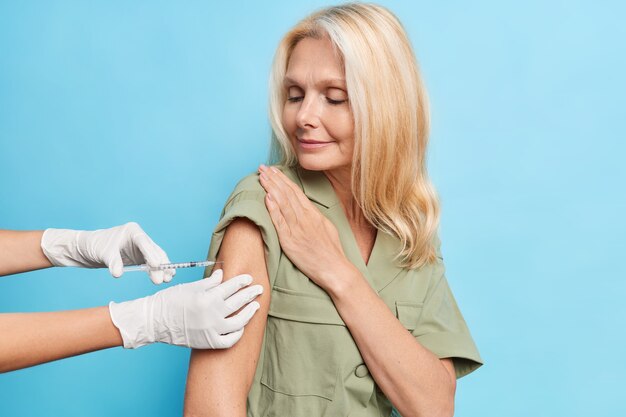 Serious middle aged woman with fair hair looks attentively at process of vaccination gets shot in shoulder to protect herself from coronavirus isolated on blue