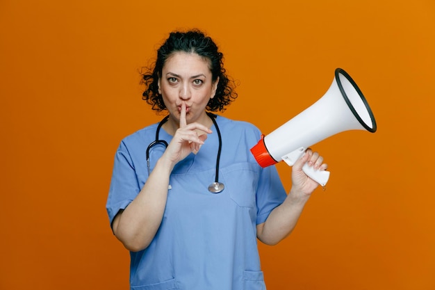 Serious middleaged female doctor wearing uniform and stethoscope around her neck holding speaker looking at camera showing silence gesture isolated on orange background