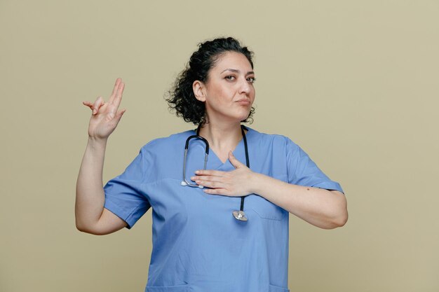 Serious middleaged female doctor wearing uniform and stethoscope around neck looking at camera making promise gesture isolated on olive background