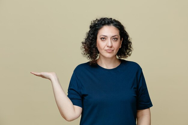 Серьезная женщина средних лет в футболке смотрит в камеру, показывая пустую руку на оливково-зеленом фоне