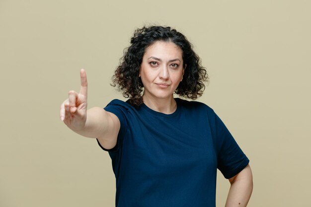 Serious middleaged woman wearing tshirt looking at camera showing one with hand isolated on olive green background