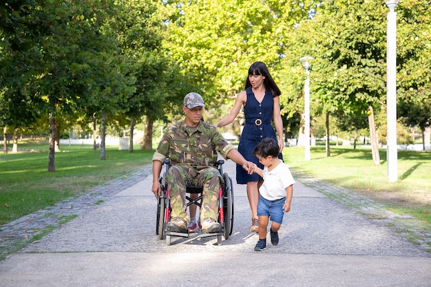 Serious military father walking in wheelchair with family. Caucasian middle-aged dad in camouflage uniform holding son hand and talking with pretty wife. Veteran of war and disability concept