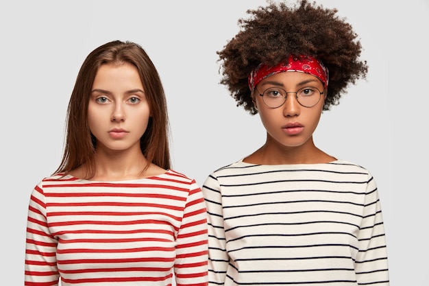 Free photo serious mixed race women sisters dressed in striped sweaters, look seriously