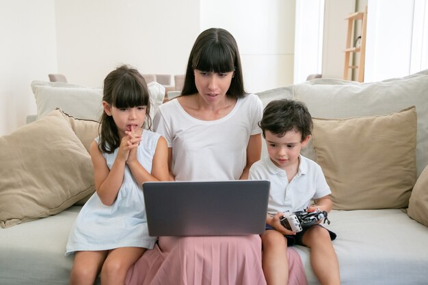Серьезная мама и двое обеспокоенных детей смотрят фильм на ноутбуке в гостиной.