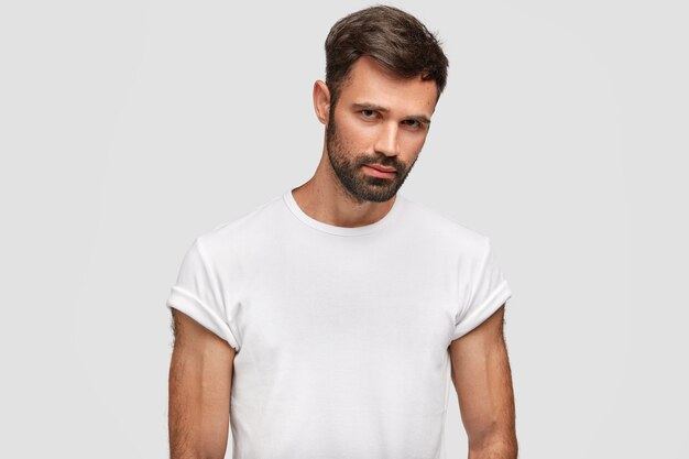 Serious muscular young male with dark stubble, hair, dressed in casual white t-shirt, has muscular body, listens attentively something, isolated over white wall. Unshaven guy stands indoor