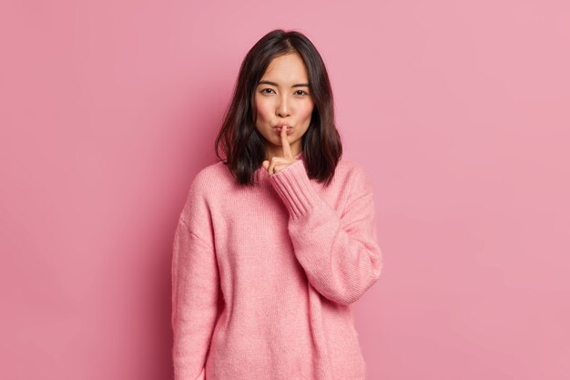 Serious mysterious brunette Asian woman presses index finger to lips makes hush gesture tells secret asks to be quiet wears long sleeved jumper poses 