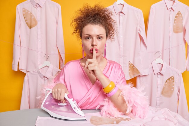 Serious mysterious housewife with curly hair makes silence gesture asks not to tell anyone she burnt shirt while ironing as has lack of experience wears dressing gown isolated over yellow wall.