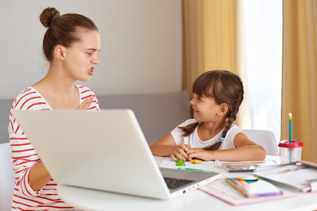 Serious nervous mother explaining home task her happy smiling daughter, woman wearing casual attire with hair ban sitting at table with schoolchild, in front of open laptop and book, online lessons.