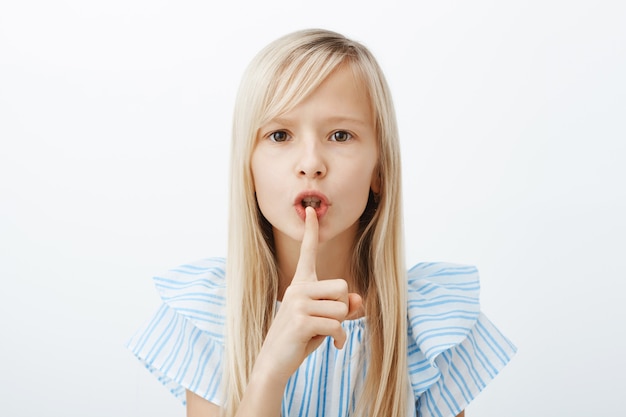 Free photo serious obedient child asks keep voice down, looking after little sister. confident adorable fair-haired young girl, bending towards, making shush sign, saying shh with index finger over mouth