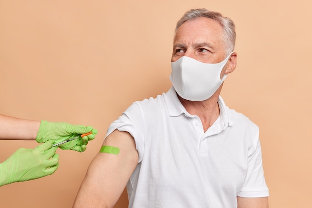 Serious old man gets vaccination against coronavirus wears protective disposable mask green adhesive tape on arm dressed in white t shirt poses near nurse who holds syringe