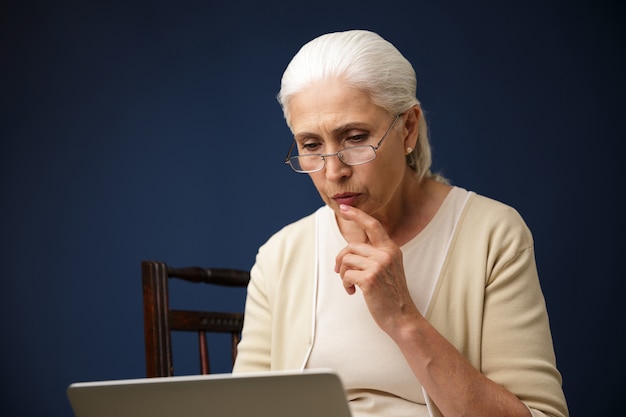 Serious old woman using laptop computer. Looking aside.