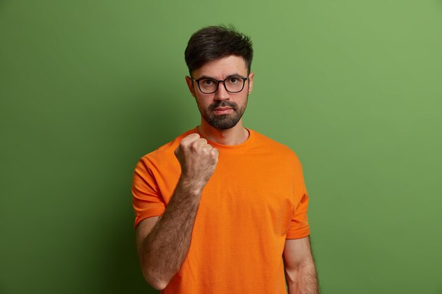 Serious outraged man shakes fist, promises to revenge, says I will show you, warns about something, looks through glasses, wears orange t shirt, expresses negative emotions, isolated on green wall