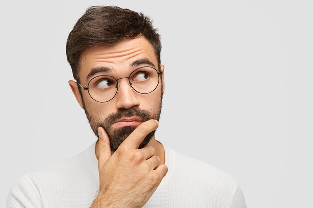 serious pensive bearded man with thick stubble, holds chin and looks pensively aside, contemplates about something while focused at upper right upper corner