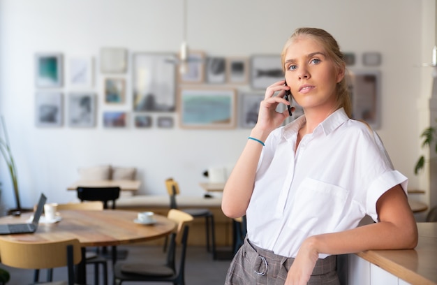 Serious pensive beautiful young woman wearing white shirt, talking on mobile phone, standing in co-working space 