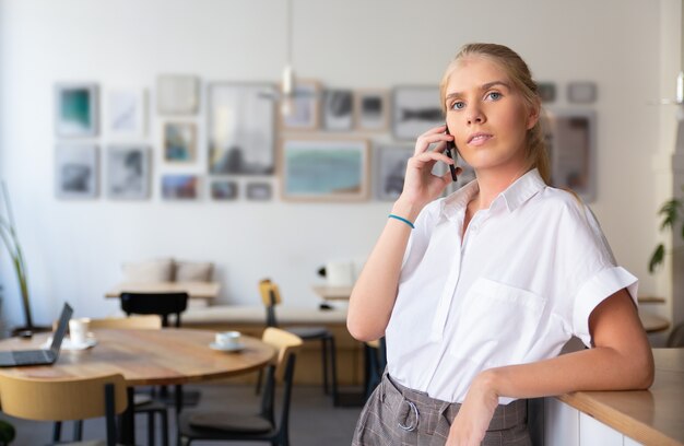 Serious pensive beautiful young woman wearing white shirt, talking on mobile phone, standing in co-working space 