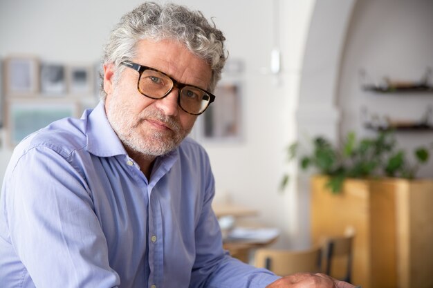 Serious pensive mature business man wearing shirt and glasses, sitting in office cafe, looking at camera