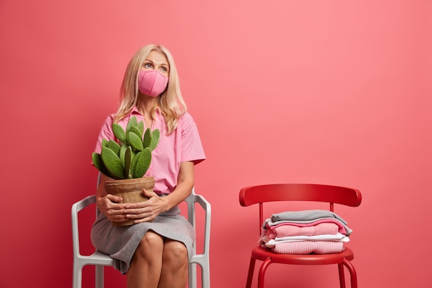 Serious pensive middle aged woman wears protective face mask thinks about safety during pandemic holds potted cactus sits on chair alone stays at home on quarantine period. Covid 19 social distancing