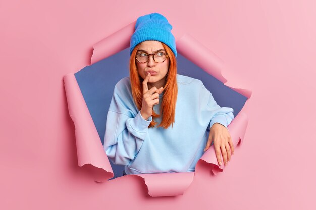 Serious pensive redhead young woman looks away with thoughtful concentrated face expression keeps index finger near lips wears blue clothing.