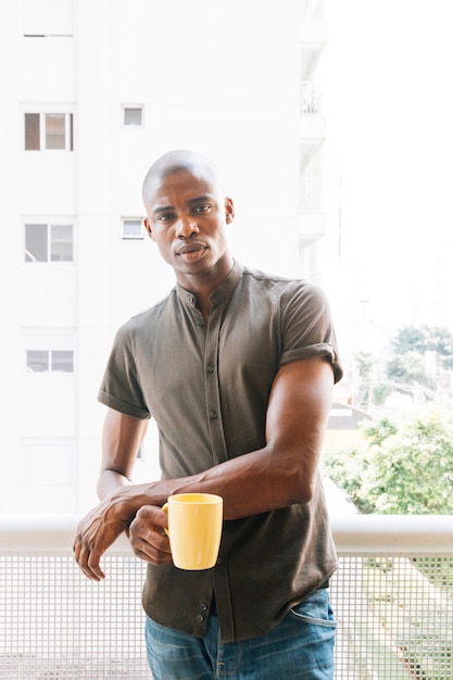 黄色のコーヒーカップを保持しているバルコニーに立っている若いアフリカ人の深刻な肖像画