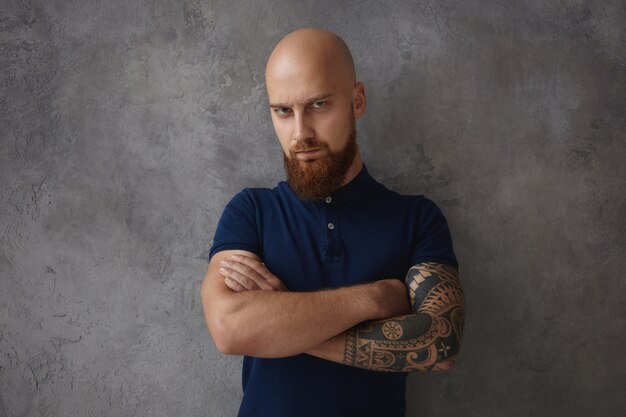 serious powerful European male with tattoo, bald head and thick beard frowning having displeased dissatisfied look, expressing his negative attitude, keeping arms folded on chest